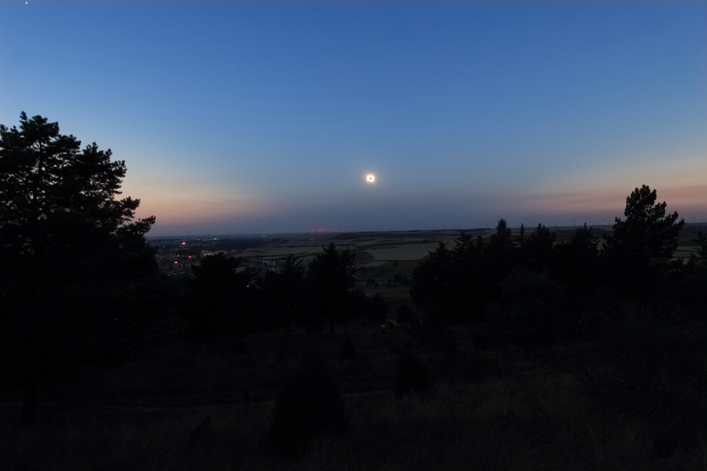Landschaft bei Burgos während der Totalität: dunkle Baumgruppen im Vordergrund, die verfinsterte Sonne als heller Punkt tief über dem Horizont, dämmeriger Himmel mit Abendrot