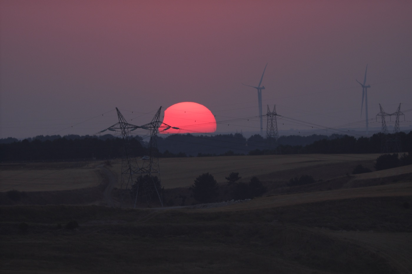 Die noch partiell verfinsterte, rot leuchtende Sonne geht über der Hochebene von Burgos unter, am Horizont Windräder und Strommasten